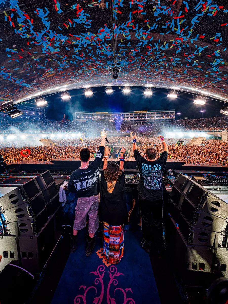 Alok entre o pai e a mãe, os DJs Swarup e Ekanta, com os braços erguidos na cabine do Mainstage do Tomorrowland, com confete caindo sobre o público