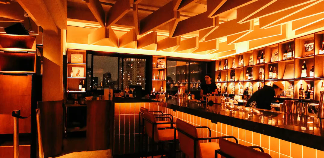 Balcão de listening bar em São Paulo, luz âmbar, prateleira de garrafas e vista da cidade pela janela